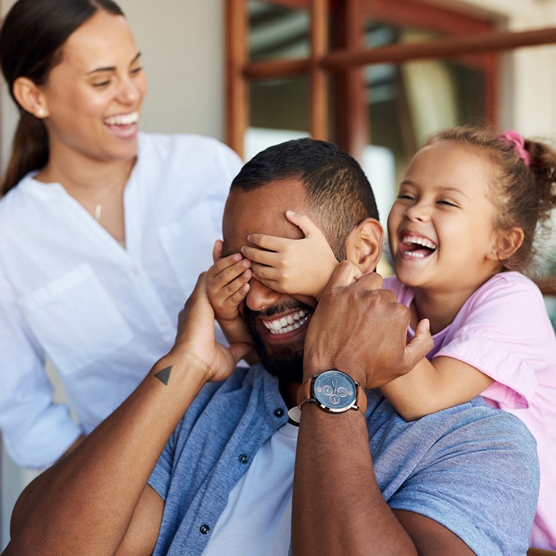 Daughter covers her fathers eyes as mother watches and laughs